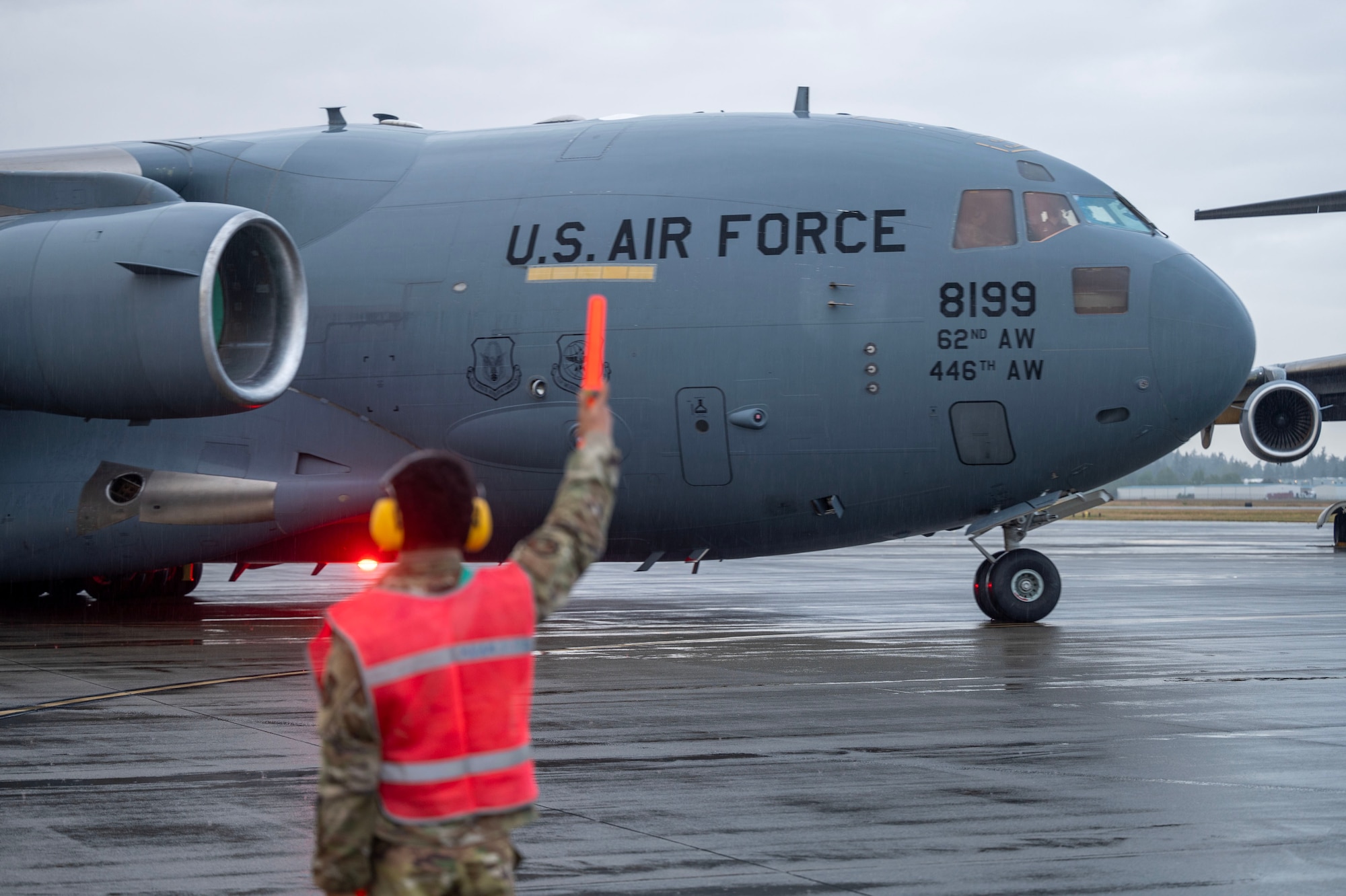 an official in a military uniform and a safety vest in the left side of the frame marshals a military plane in the right side of the frame