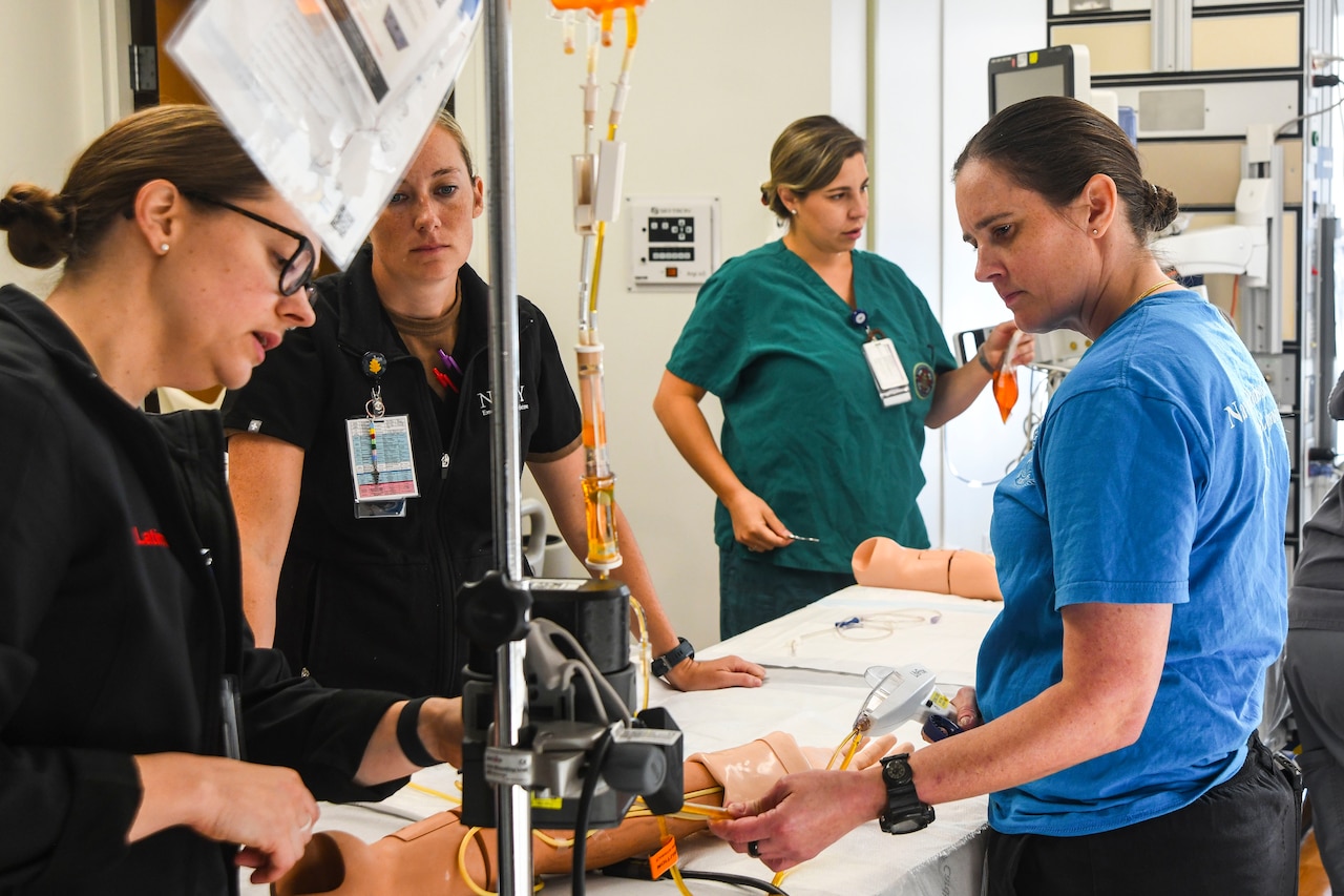 Four women wearing medical attire stand over an IV training arm with medical equipment attached; one provides instruction while the others observe.