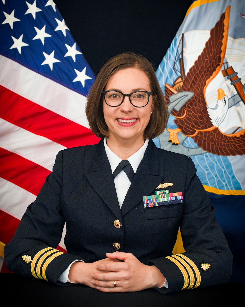 A woman wearing a formal military uniform poses for a portrait with an American flag and a Navy flag in the background.