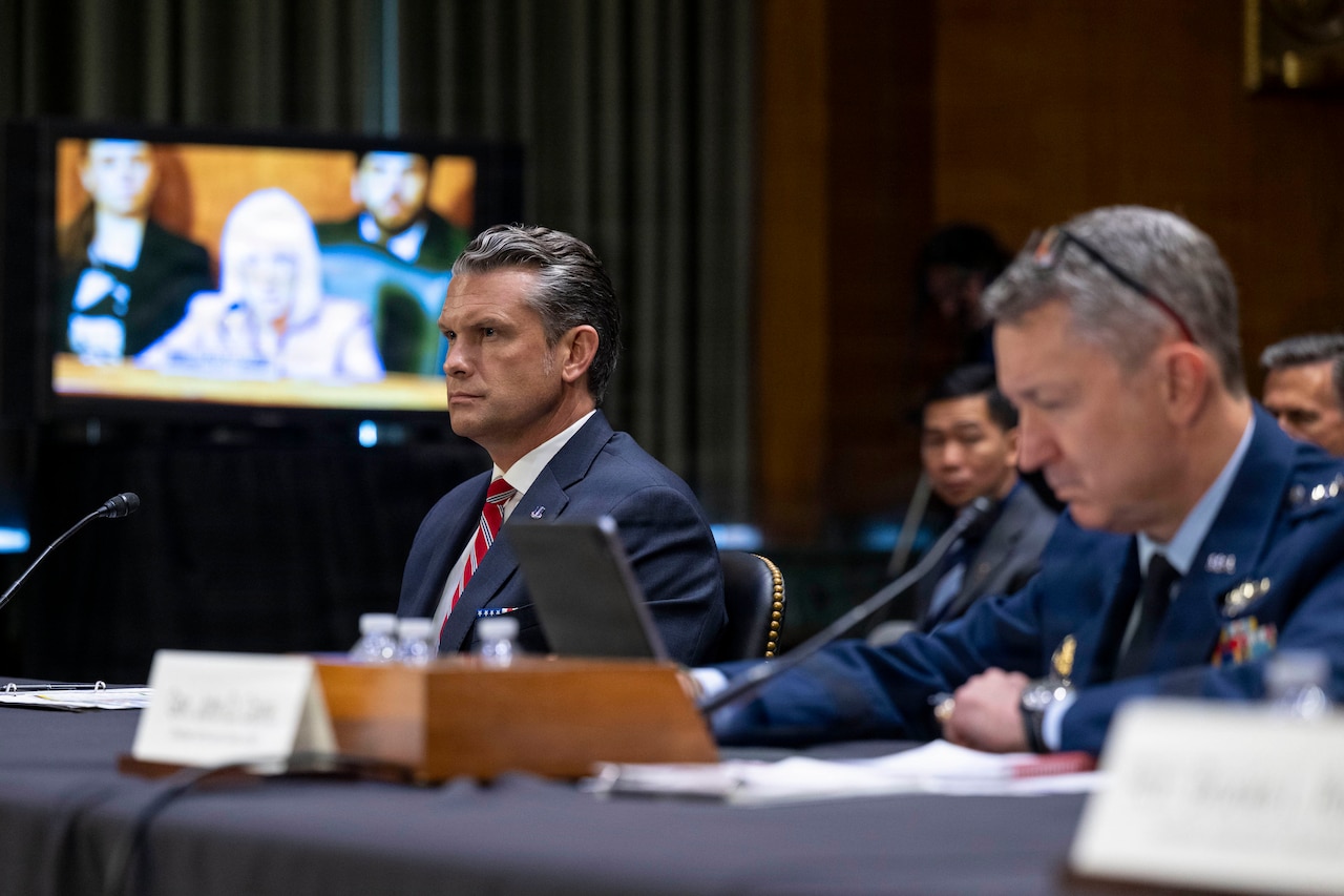 Secretary of War Pete Hegseth and Air Force Gen. Dan Caine sit in front of microphones at a table.