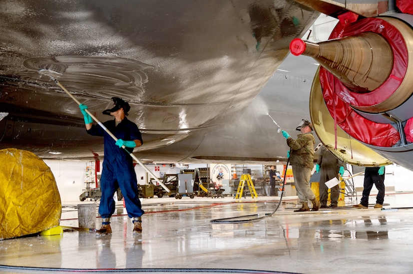 Two people wearing military jumpsuits, clean the underside of a military jet.