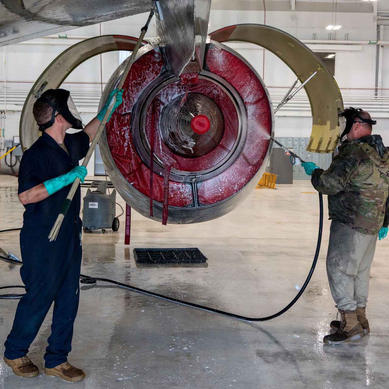Two men clean the engine area of a military jet. One man is wearing a blue jumpsuit and the other is wearing a camouflage military jacket.