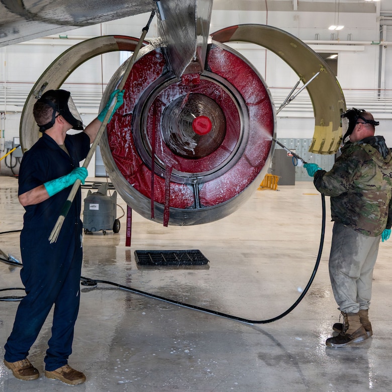 Two men clean the engine area of a military jet. One man is wearing a blue jumpsuit and the other is wearing a camouflage military jacket.