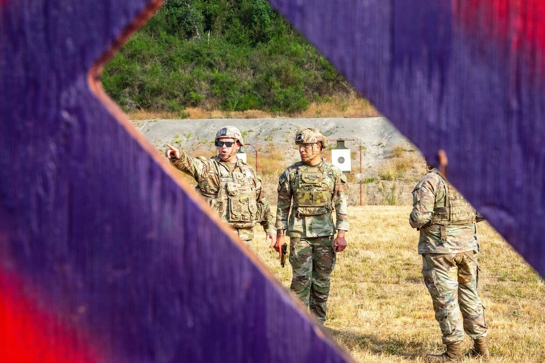 Soldiers talk to each other in a field while standing in front of targets as through a window in a colorful wooden structure.