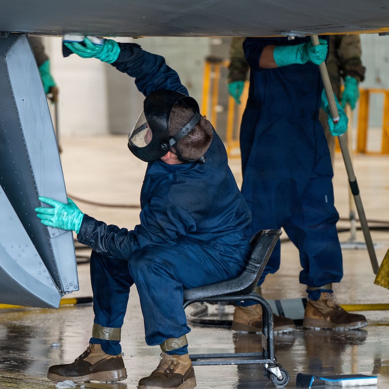 A man wearing face protection and a blue jumpsuit, cleans the bottom of a military jet.