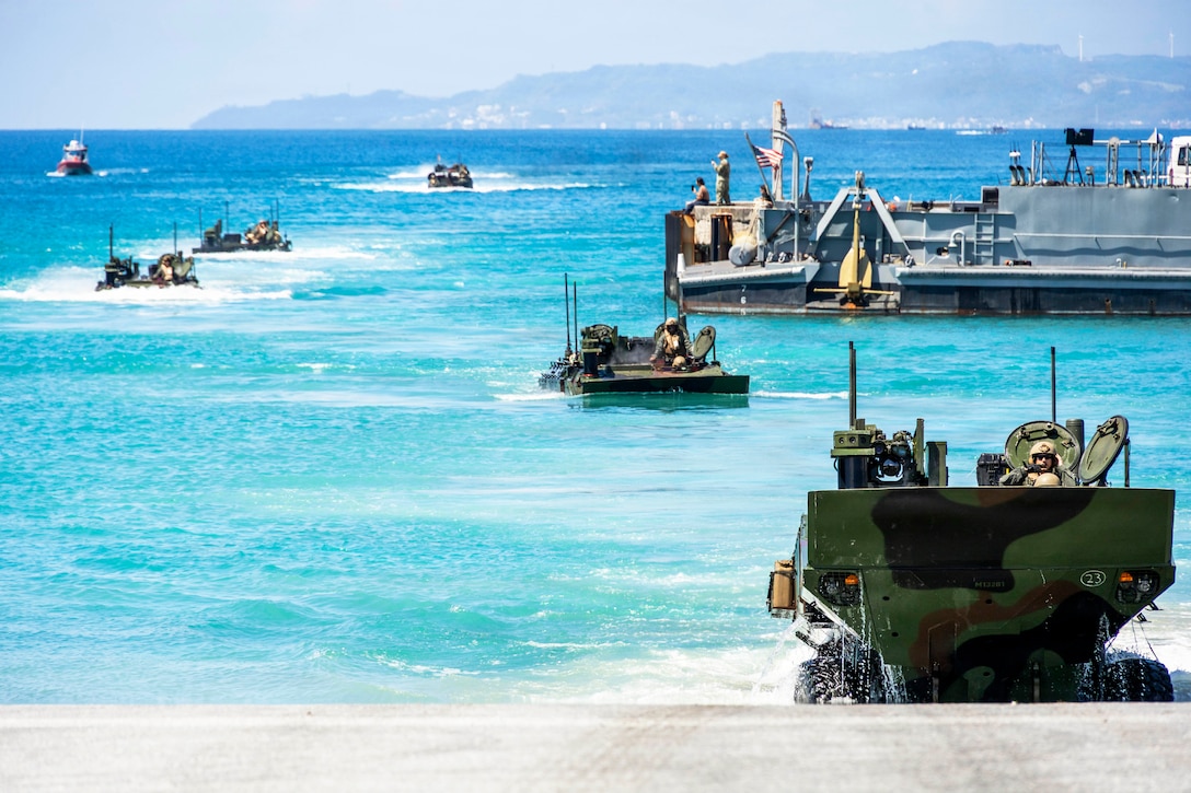 Amphibious combat vehicles transit light blue waters near a ship during the day.