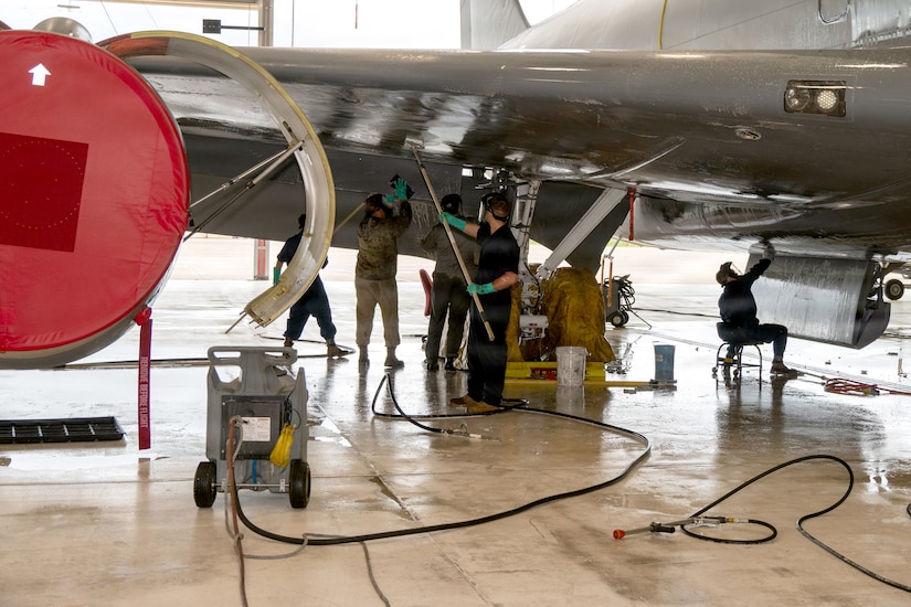 Five people wearing military jumpsuits, clean the underside of a military jet.