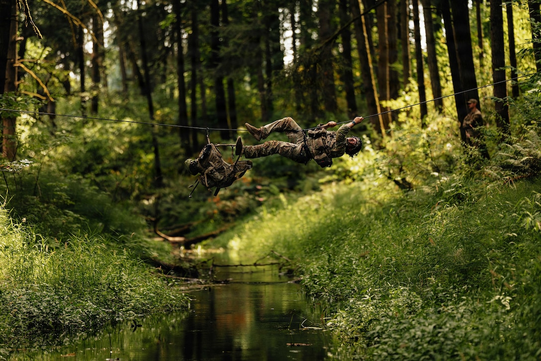 A soldier in a camouflage uniform horizontally climbs across a rope over a creek in a wooded area.