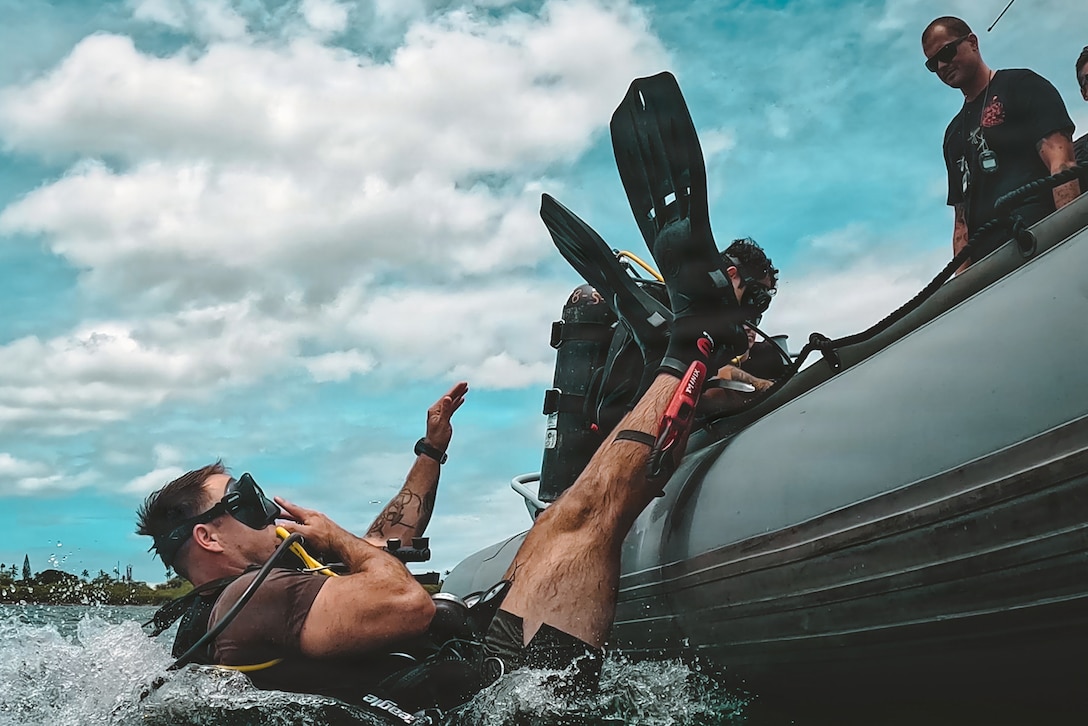 A sailor wearing a brown T-shirt and green shorts, goggles and a snorkel enters water back first with flippers in the air. Other sailors look on, and one prepares to plunge in.