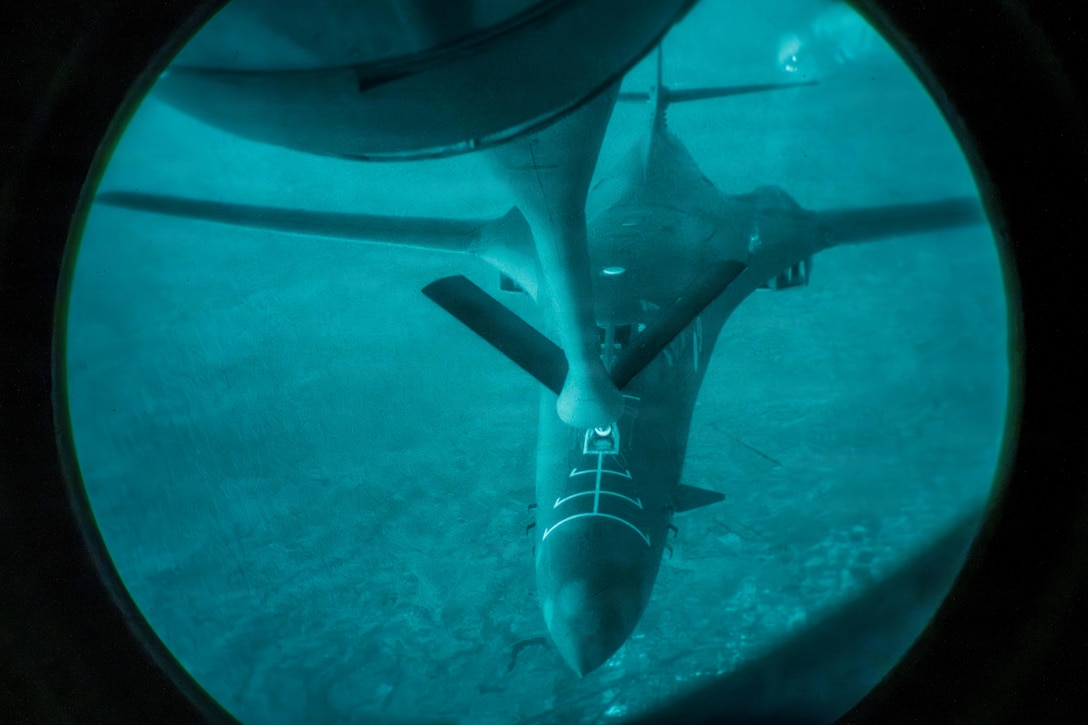 A jet refuels in the air from the boom of a partially visible aircraft above it. The image is circular, with a blue tint.