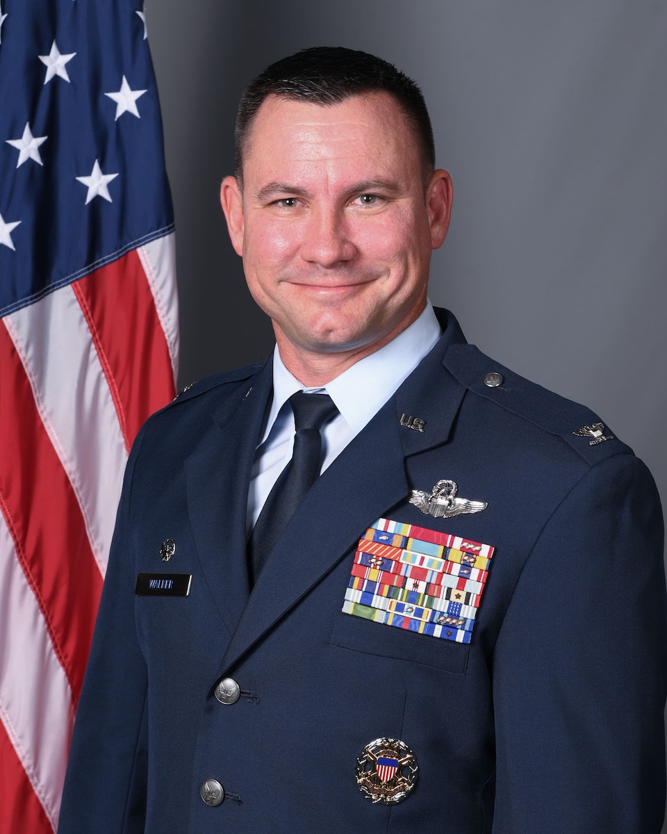 An Air Force Colonel poses for an official photo with a gray background and an American flag to his left.