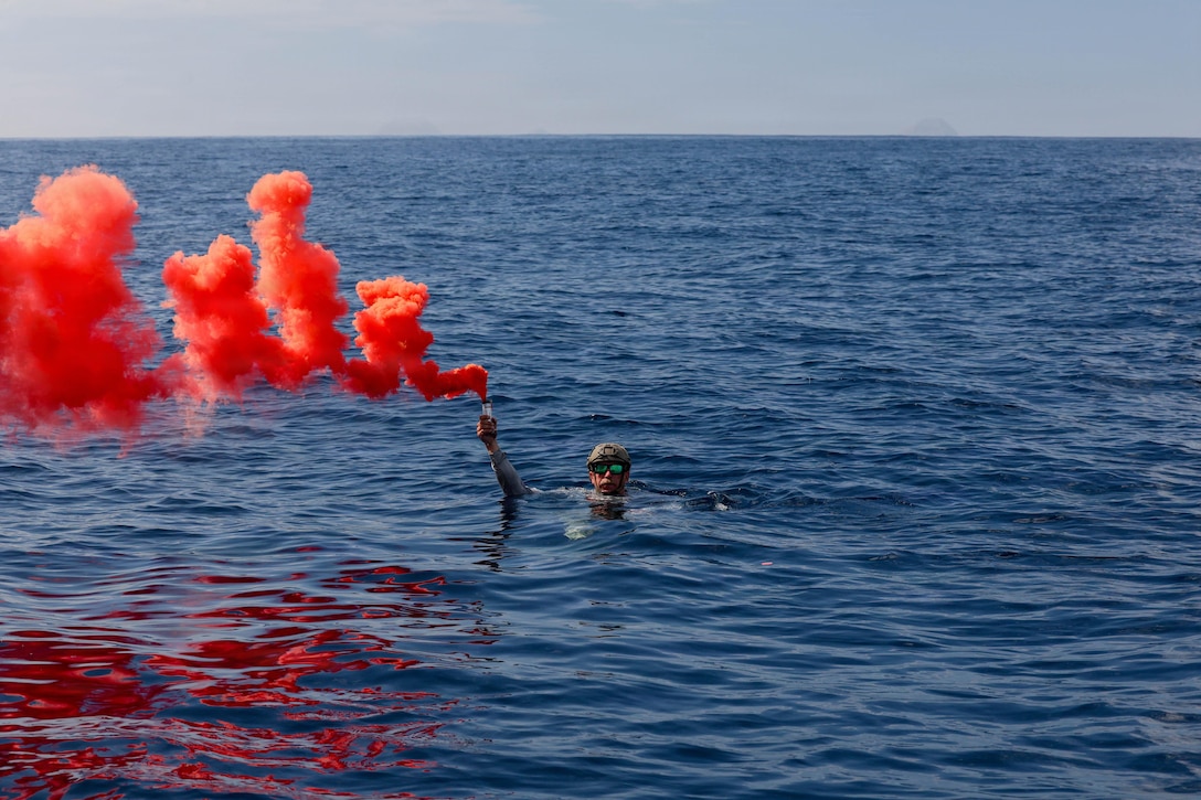 A sailor in a helmet and sunglasses has their head above water in the ocean and holds up a flare with red smoke coming out of it with one hand.