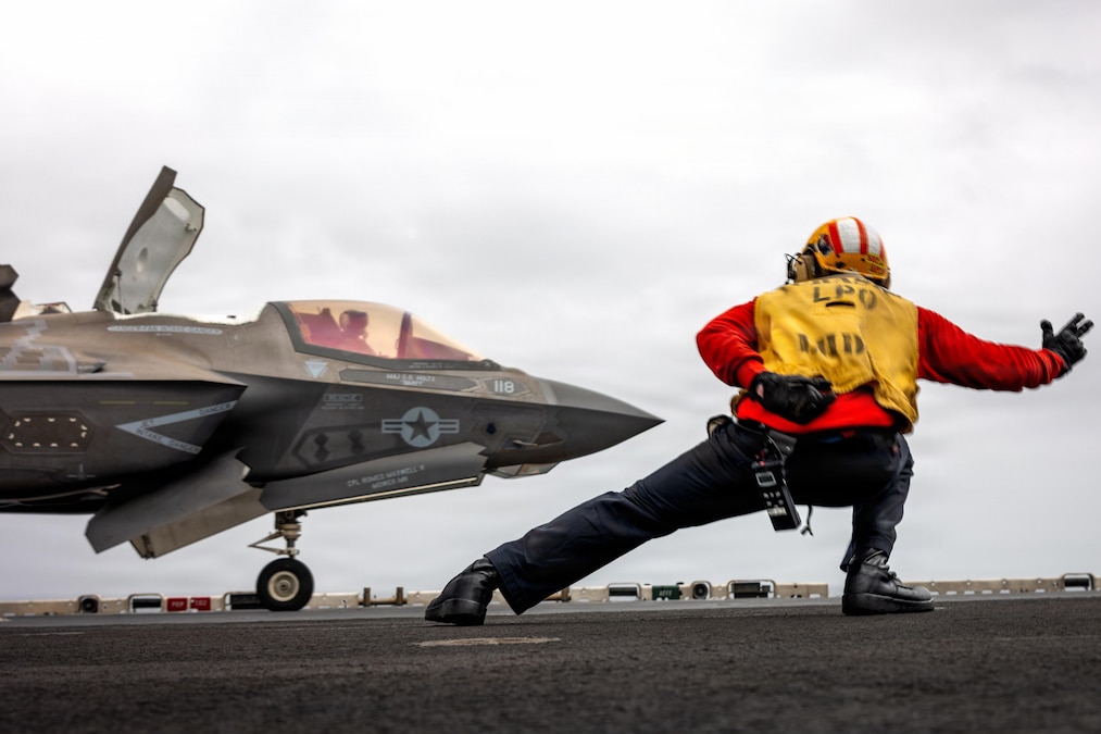 A sailor in a yellow vest over clothes lunges and holds up a thumb and two fingers while looking at a pilot who is in a fighter jet on the deck of a ship.