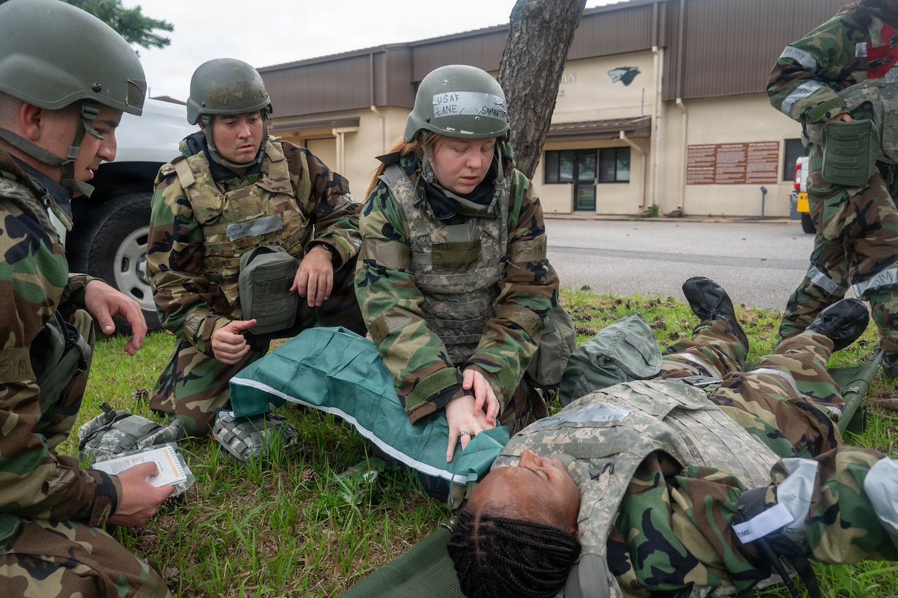 A woman in a camouflage military uniform and helmet checks on a simulated patient in similar attire lying on the ground.
