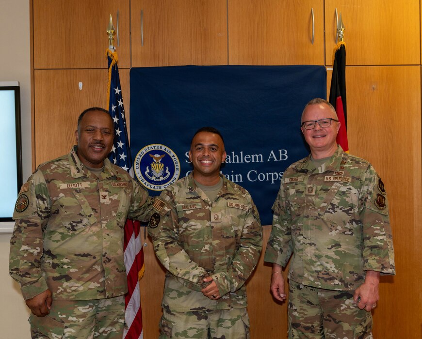 U.S. Air Force Col. Ralph Elliott, left, and Senior Master Sgt. Andrew J. Fraser, right, stand with U.S. Air Force Master Sgt. Eliseo Malave, Military and Family Readiness Center readiness senior noncommissioned officer, after presenting Malave with a commander's coin at Spangdahlem Air Base, Germany, June 24, 2026. Malave was recognized for his outstanding performance and continued support to the chapel community at Spangdahlem. Elliott is the Europe-Air Forces Africa command chaplain, and Fraser is the Air National Guard liaison to Headquarters USAFE-AFAFRICA Office of the Command Chaplain senior enlisted leader. (U.S. Air Force photo by Airman 1st Class Kacie Ramos)