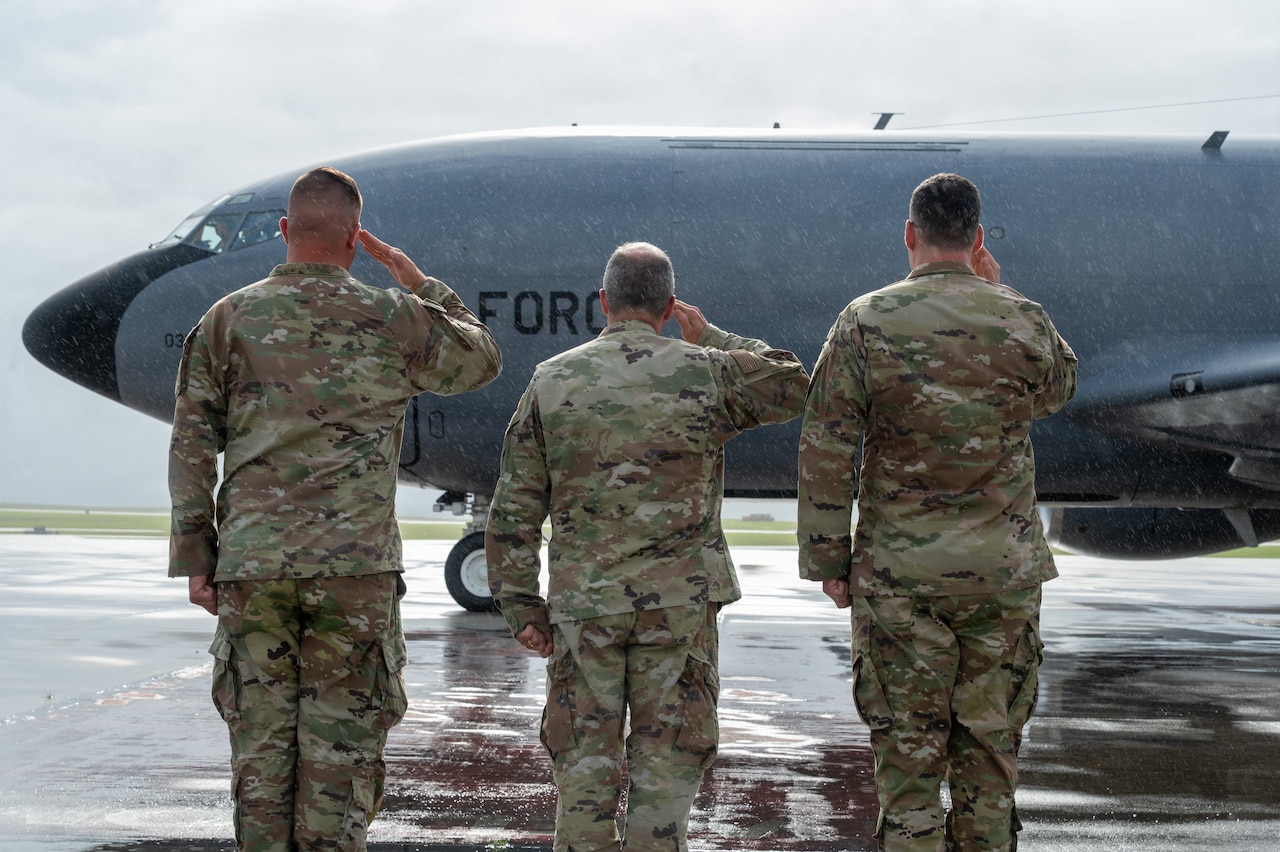 Three men wearing camouflage military uniforms salute a military aircraft.