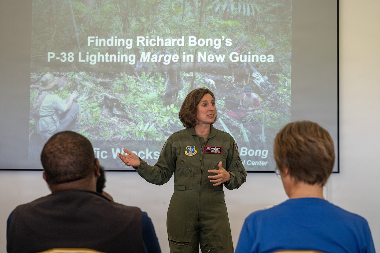 A woman dressed in an Air Force flight suit stands in front of a projector screen to address a group of seated people.