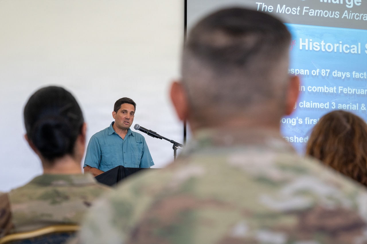 A man wearing a blue shirt stands at a lectern and addresses a crowd.