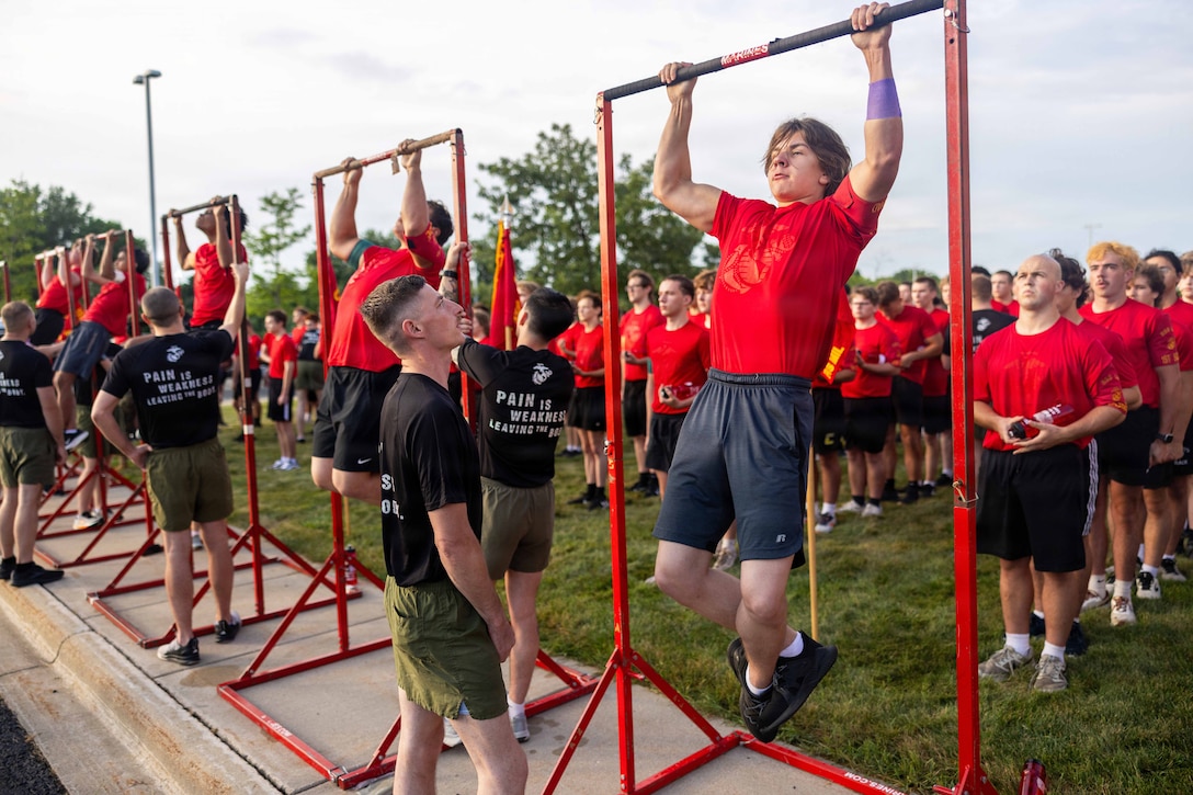 Marine Corps poolees in red shirts conduct outdoor pullups while being evaluated as dozens of poolees stand in a field waiting for their turn.