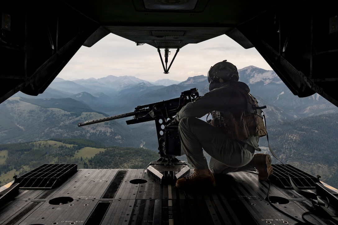 A Marine kneels in front of a weapon on the open doorway of an airborne helicopter with mountains in the background.