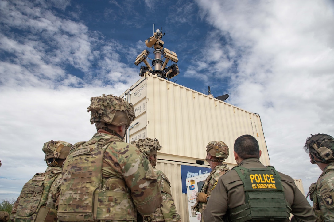 Soldiers and a Border Patrol agent in tactical gear look up at a motion sensor array positioned on stacked containers against a blue sky.