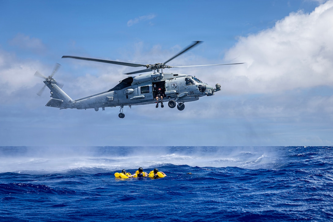 A few service members wearing yellow floatation devices float in a royal blue ocean. Another service member wearing flippers sits in a helicopter above them.
