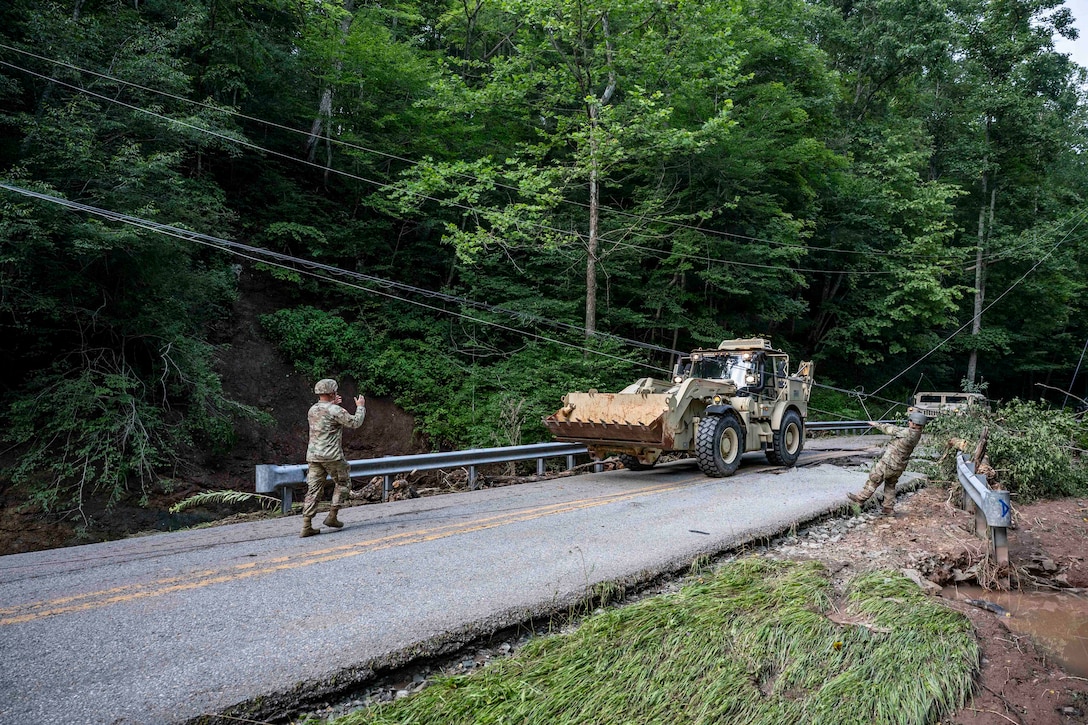 Two guardsmen direct someone driving a bulldozer on a two-lane road to help the driver get past downed electrical wires. Trees and mud surround the road.