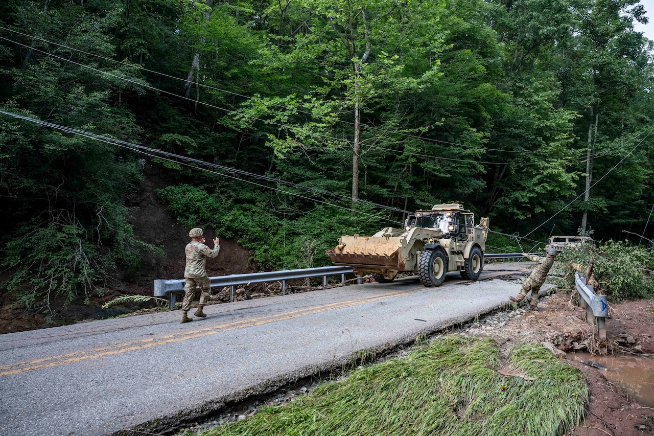 Two people in camouflage military uniforms direct the driver of a bulldozer on a two-lane road; trees and mud surround the road.