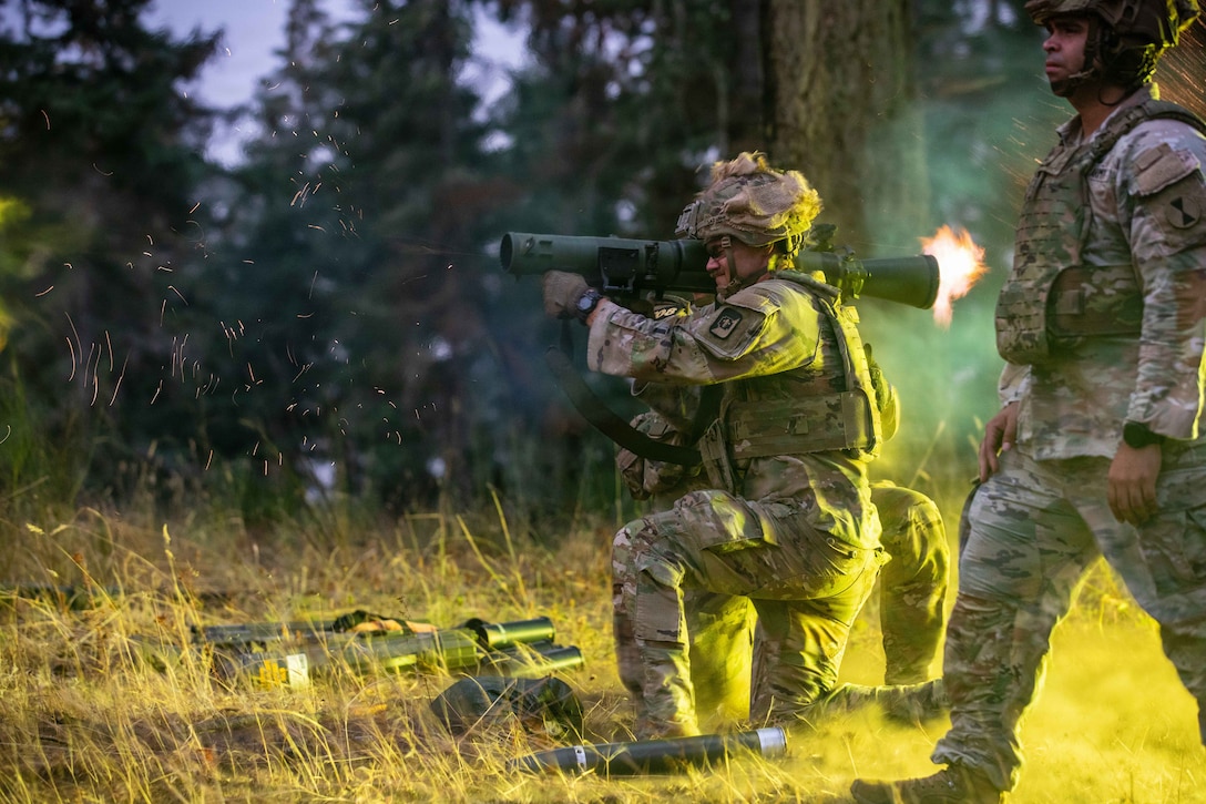 A soldier kneeling on a sunlit field fires a recoilless rifle, and fire shoots out the back of the weapon. Two other soldiers are nearby and weapons are on the ground.
