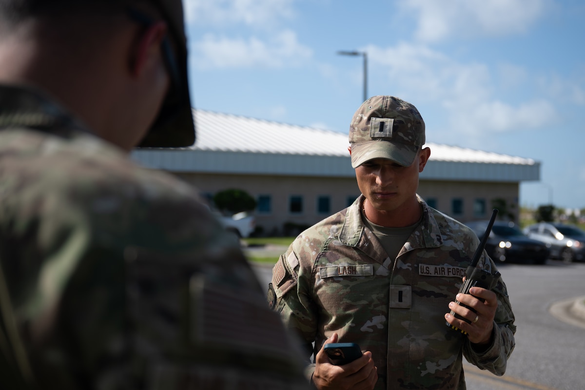 An Airman in a camouflage uniform and patrol cap looks down at his phone while holding a radio, standing outdoors and conversing with another service member.