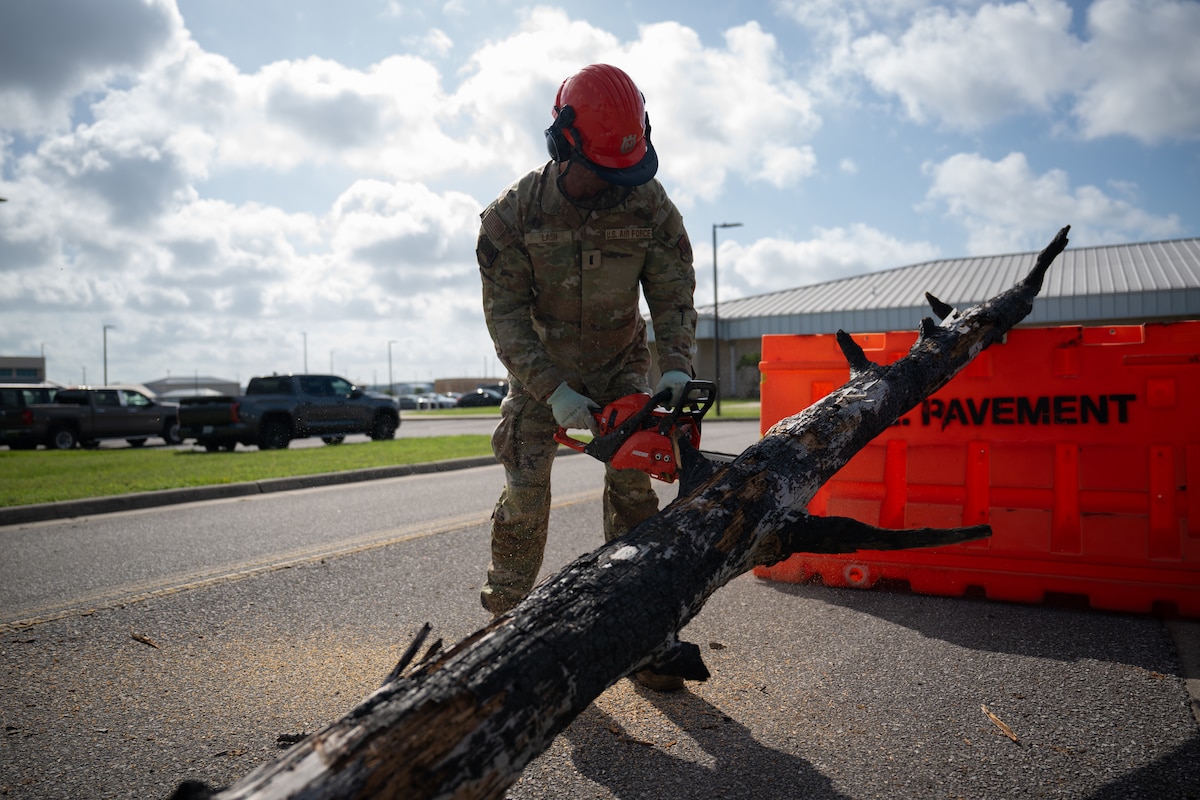 An Airman wearing a red hard hat and protective gear uses a chainsaw to cut a large, burnt tree limb lying on a paved road.