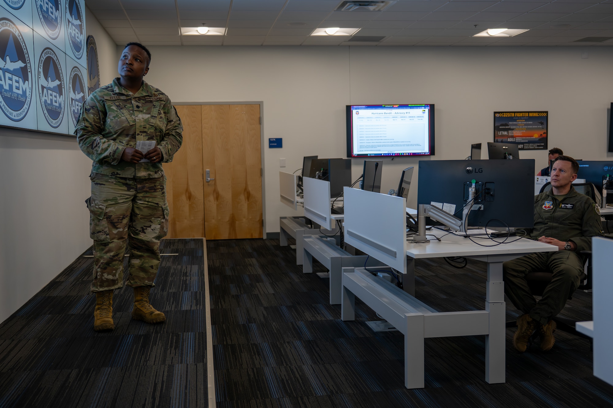 A female Airman in camouflage uniform stands and briefs a group in a modern training room, while a pilot in a flight suit listens from his desk. A large screen with Air Force Emergency Management logos is on the wall.