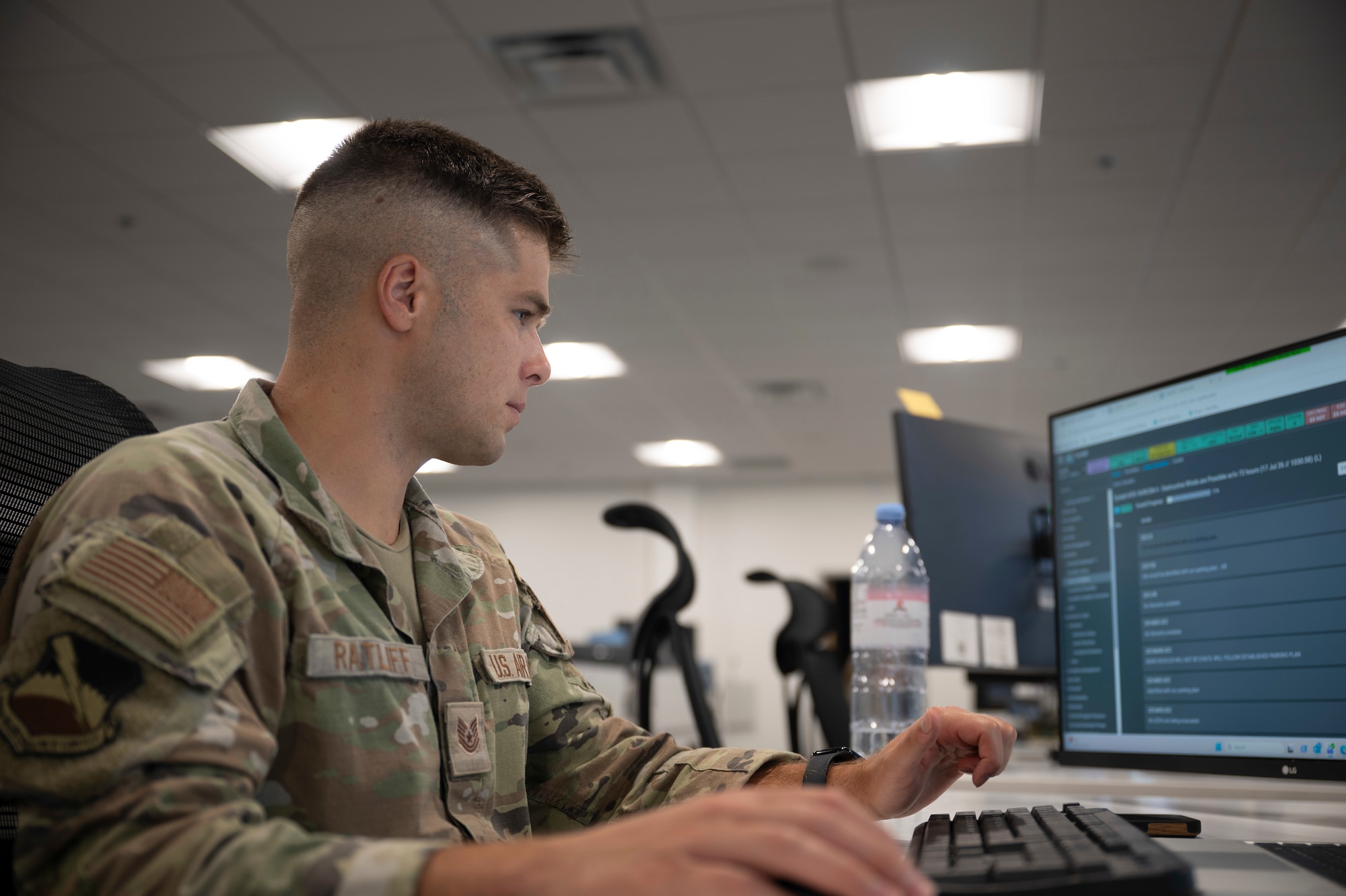 A uniformed Airman with a high-and-tight haircut sits at a computer, focused on the screen while using a keyboard and mouse in an office environment.