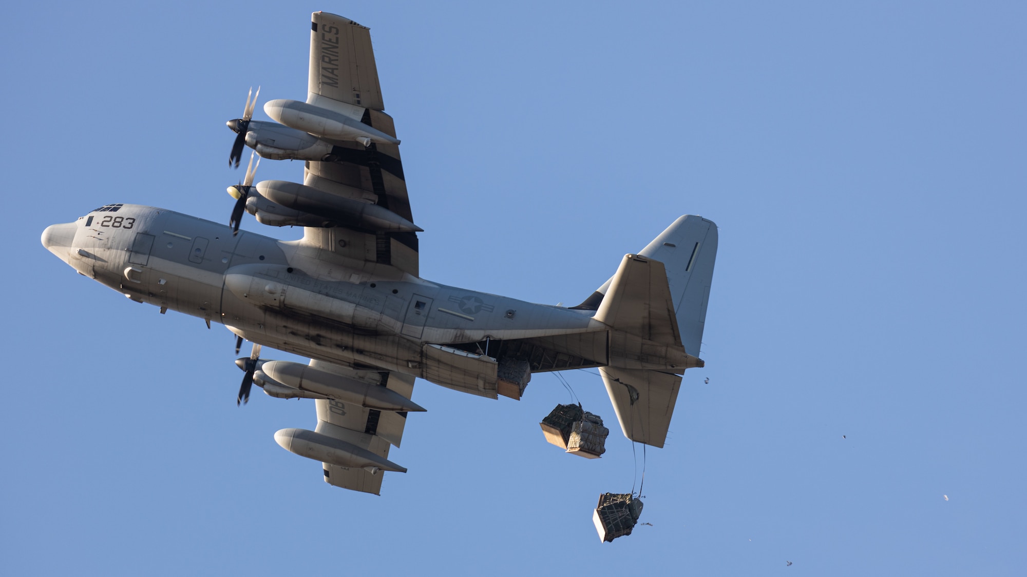 Grey aircraft flying in the sky while dropping cargo out the back of the plane.