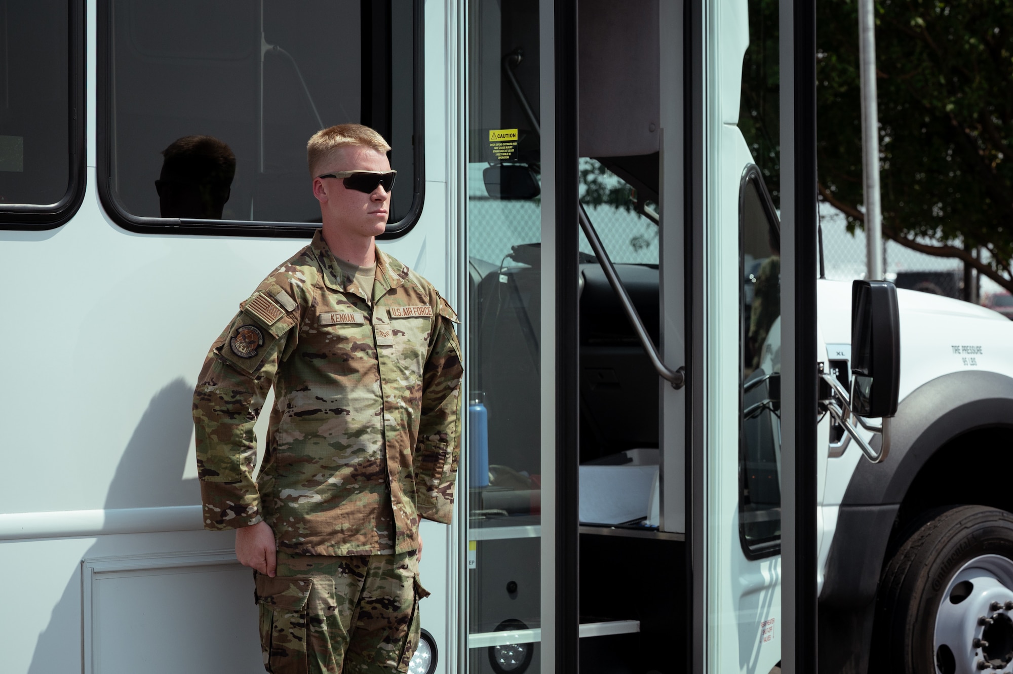A man in military camouflage uniform stands next to a white bus at the position of attention.