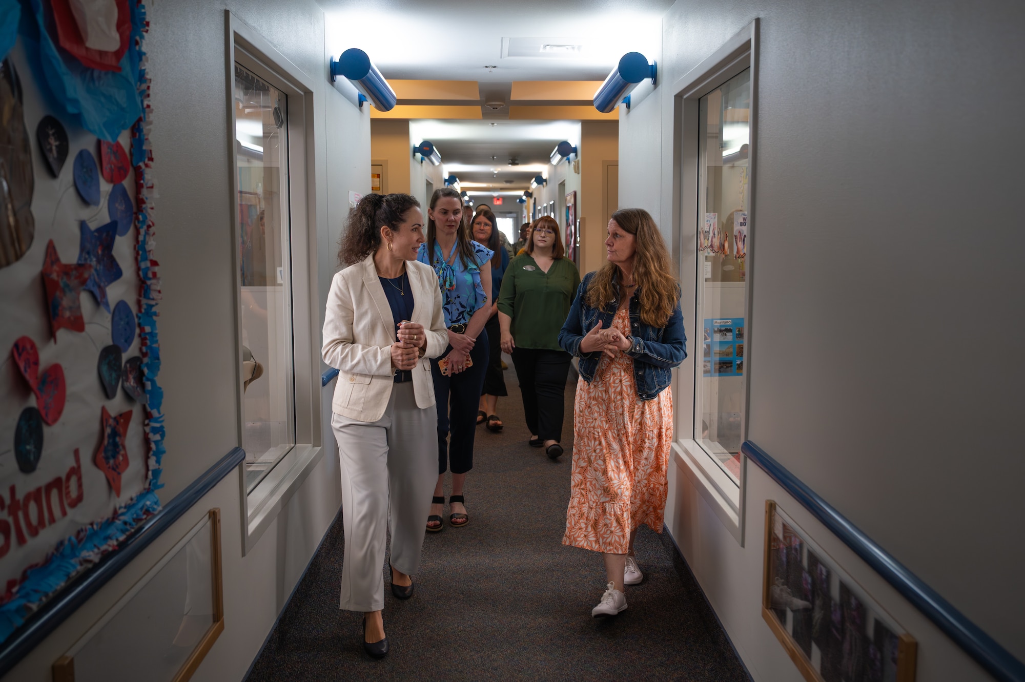 Two women speak with each other as they walk down the center of a hallway toward the camera.