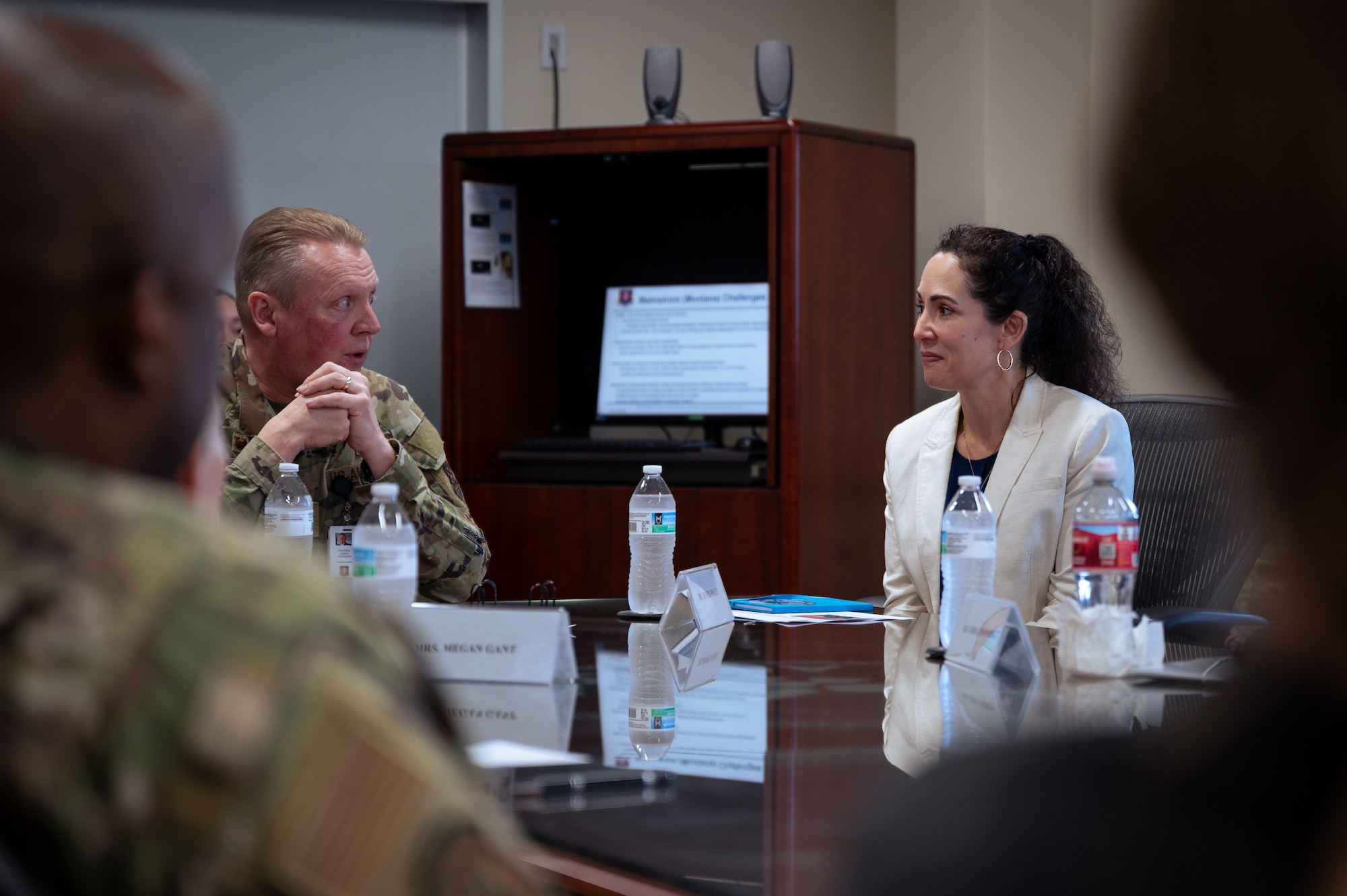 A woman with black hair in a ponytail and dressed in a white blazer sits at a table across from a man wearing a military camouflage uniform as they talk.