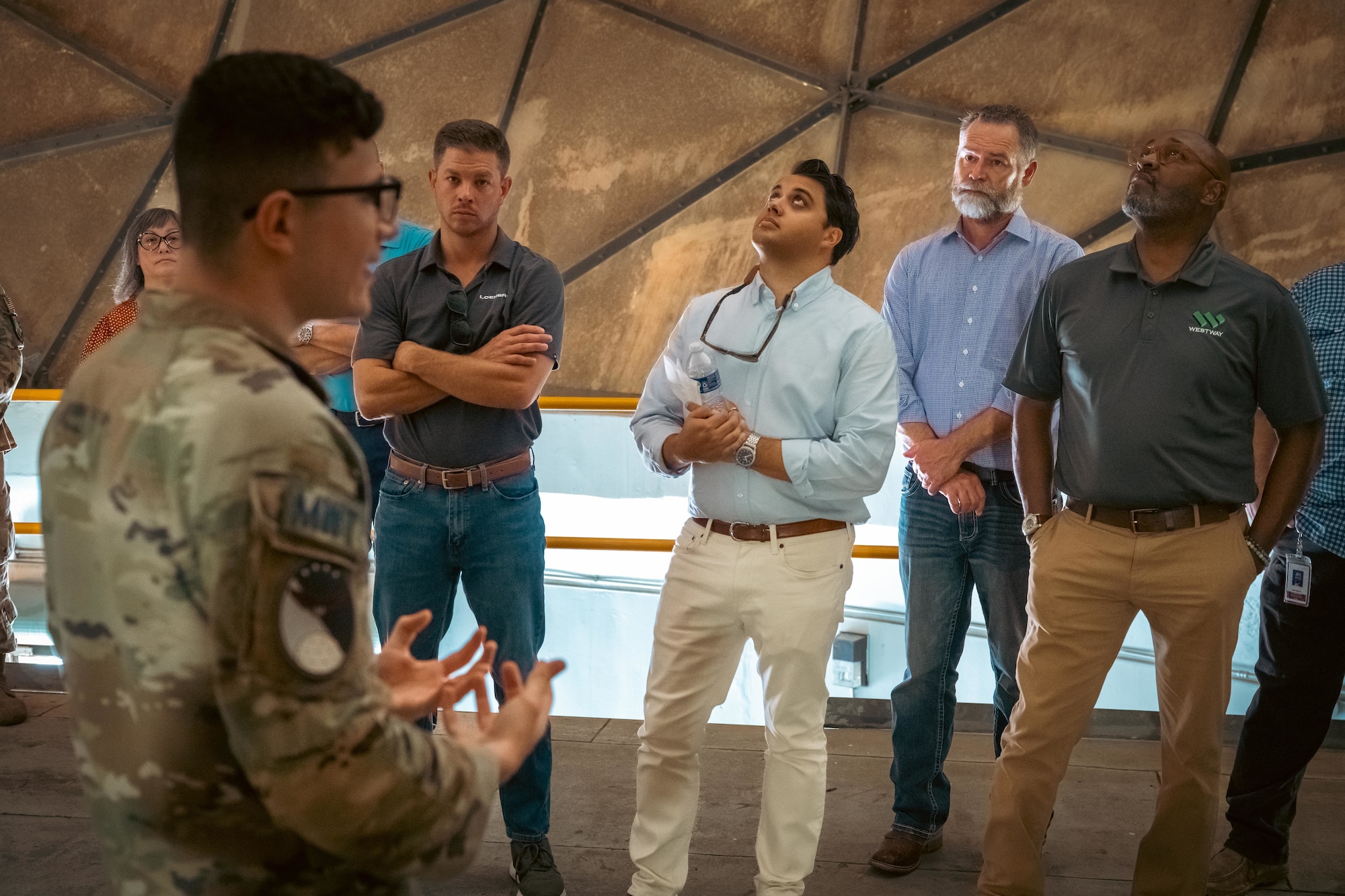 6 people stand in attendance to a briefing by a uniformed service member