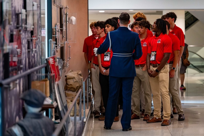 A rear-facing man wearing a formal military uniform gives a tour to a group of boys in a hallway.