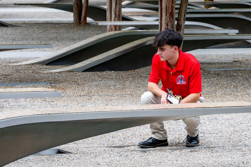 A boy wearing casual attire crouches near a bench to read a plaque outside at a memorial.