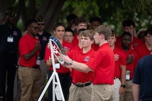 Two boys wearing casual attire place a wreath on a stand outside as more than a dozen other boys in similar attire observe.