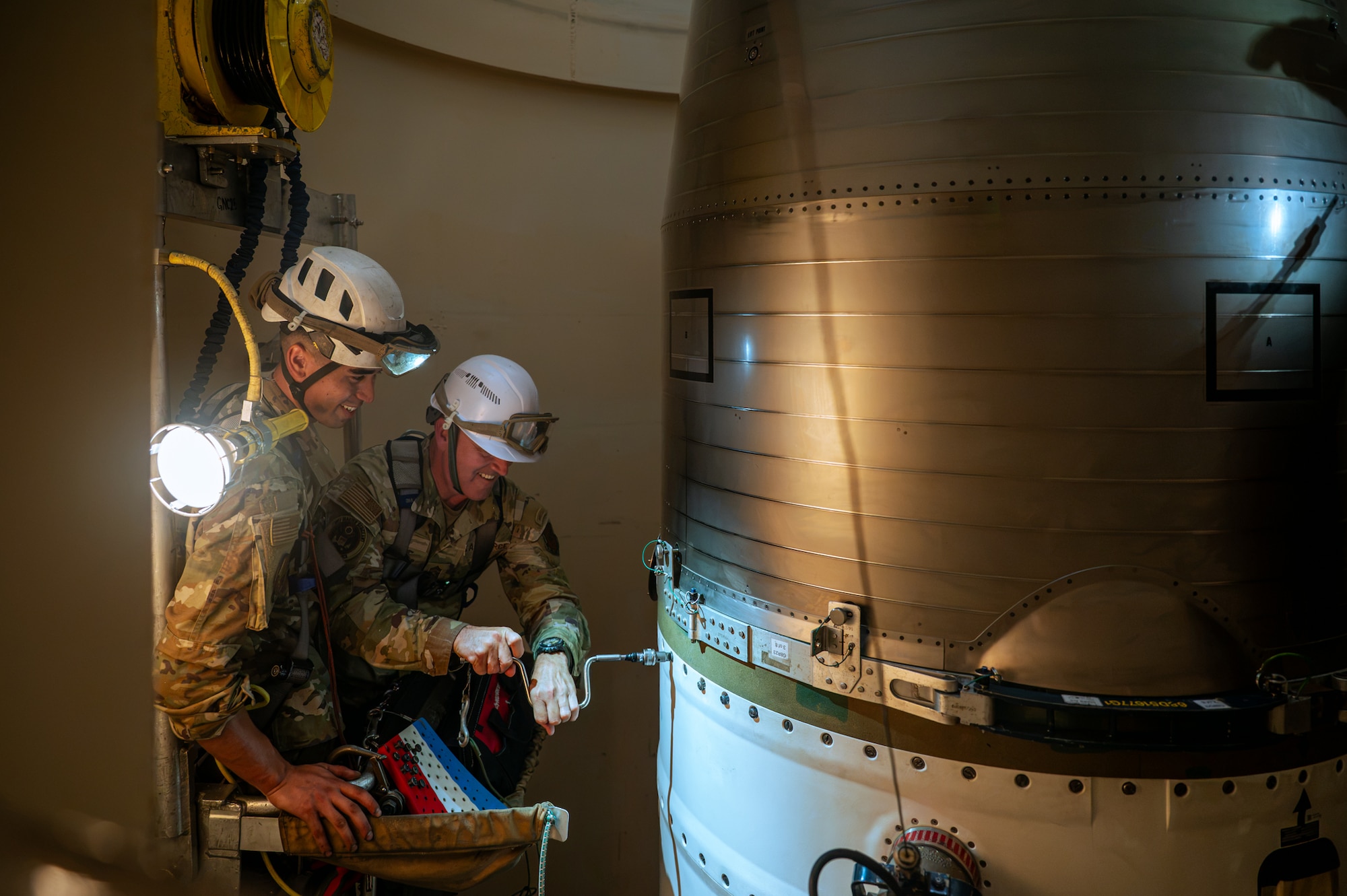Two men in military camouflage uniforms and white hard hats work together inside of a missile silo.