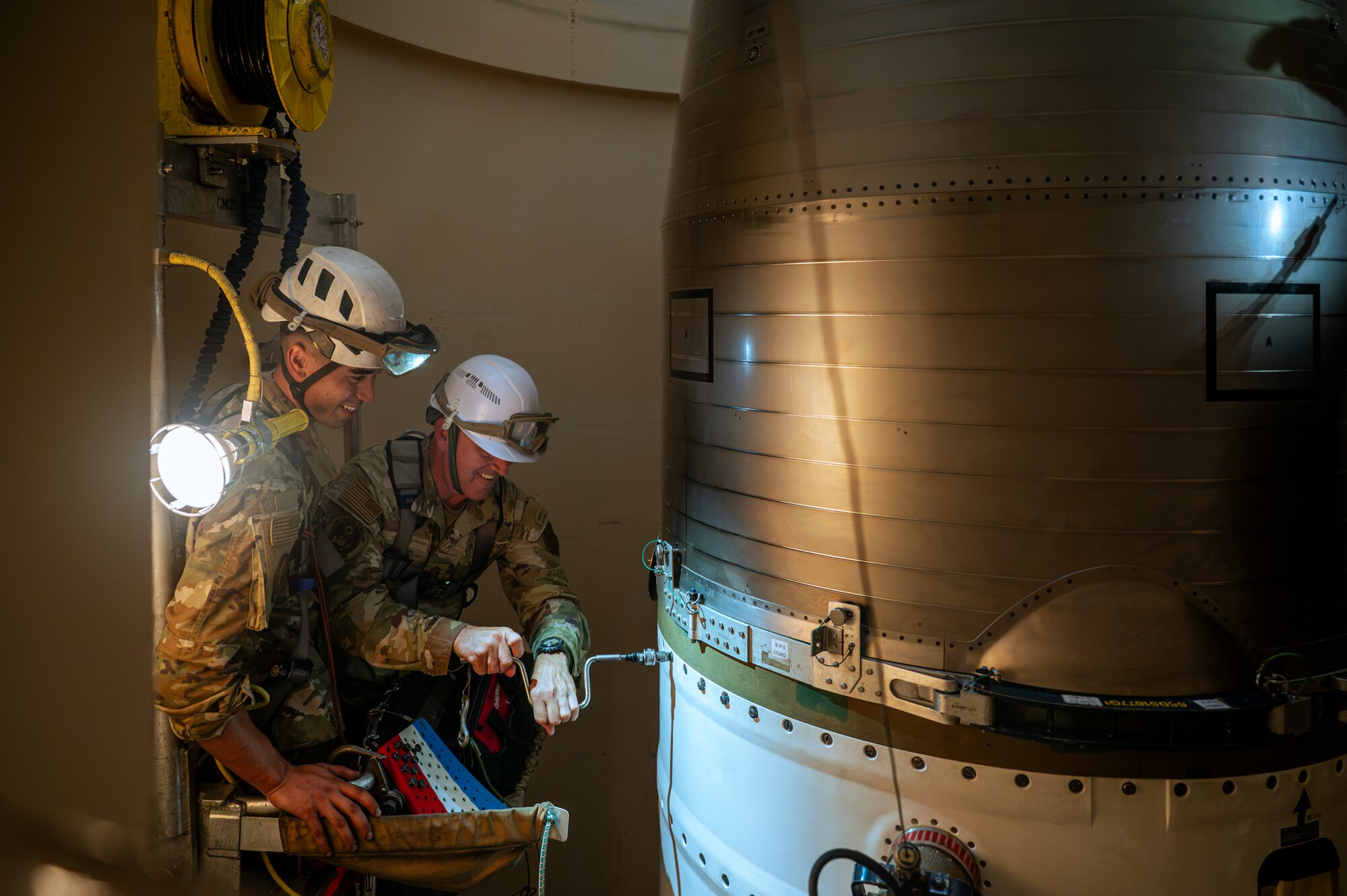 Two men in military camouflage uniforms and white hard hats work together inside of a missile silo.
