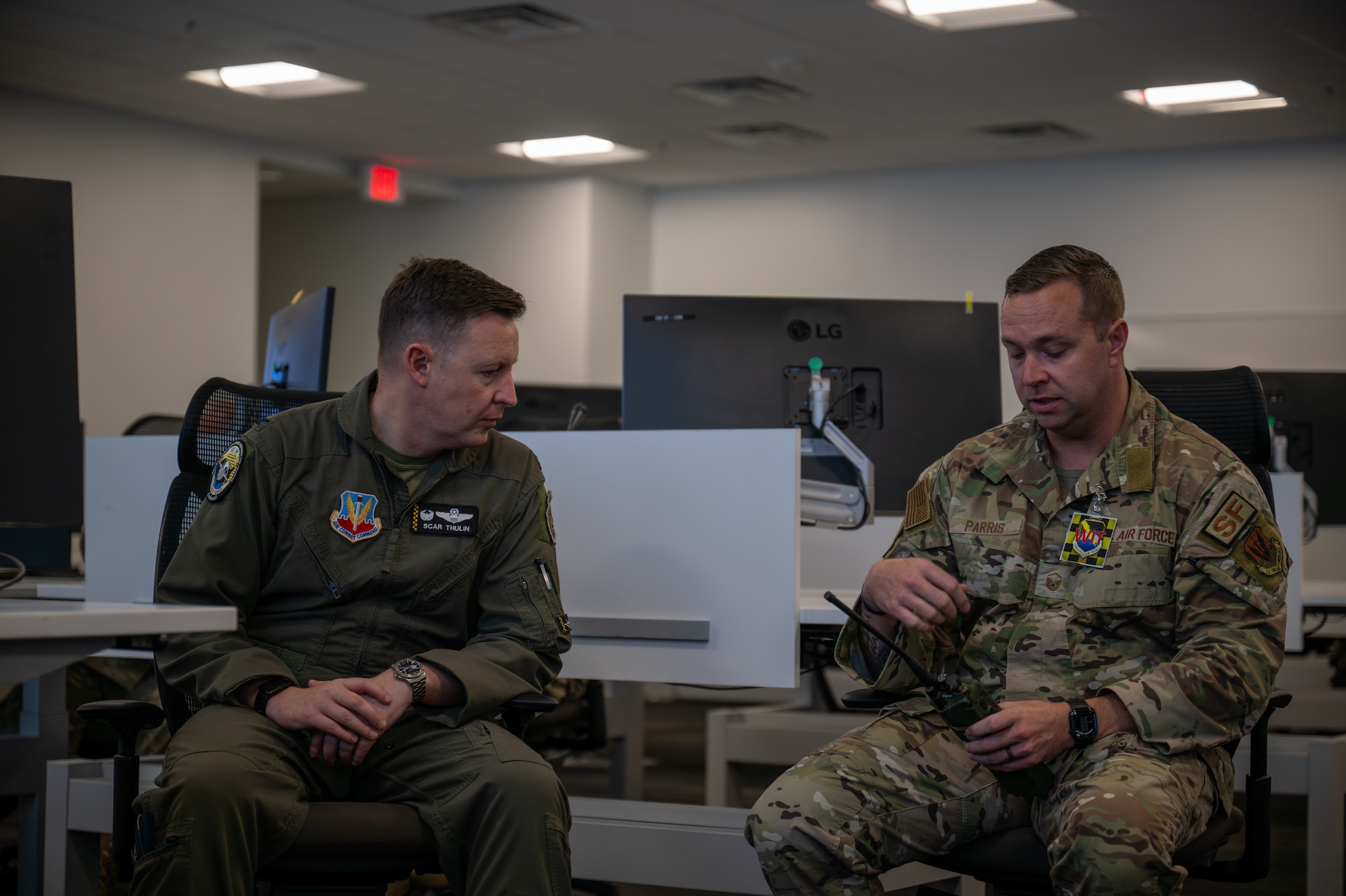 Two male Airmen, one in a flight suit and one in a camouflage uniform, are seated and engaged in a serious conversation in an office setting. The Airman in camouflage is holding a radio.