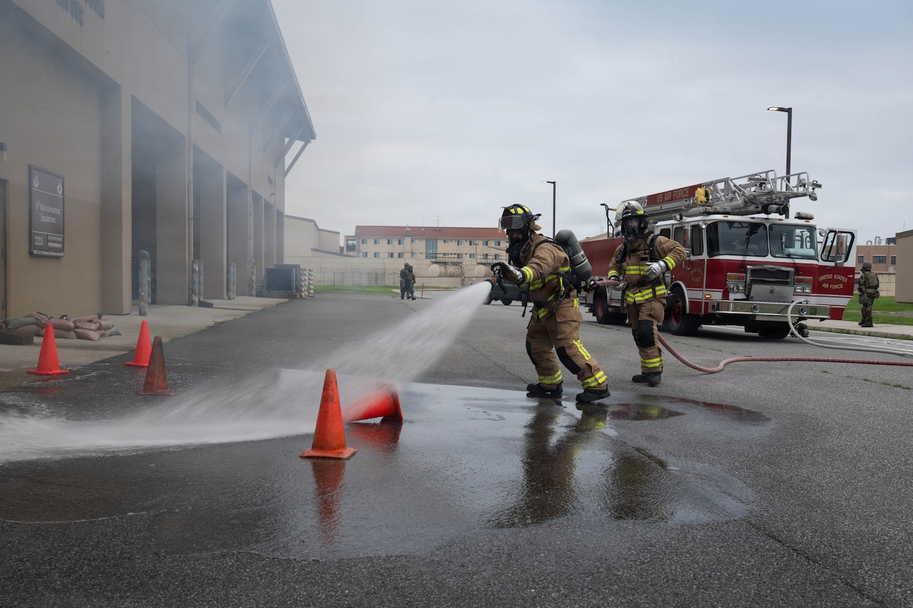 A person in a firefighter uniform uses a hose to spray water outside a building as another person in similar attire holds the hose.