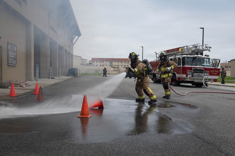 A person in a firefighter uniform uses a hose to spray water outside a building as another person in similar attire holds the hose.