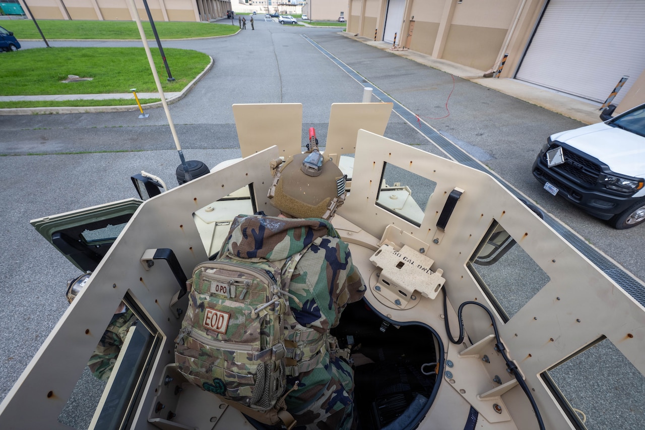 A person in a camouflage military uniform aims a rifle while on top of a military vehicle.