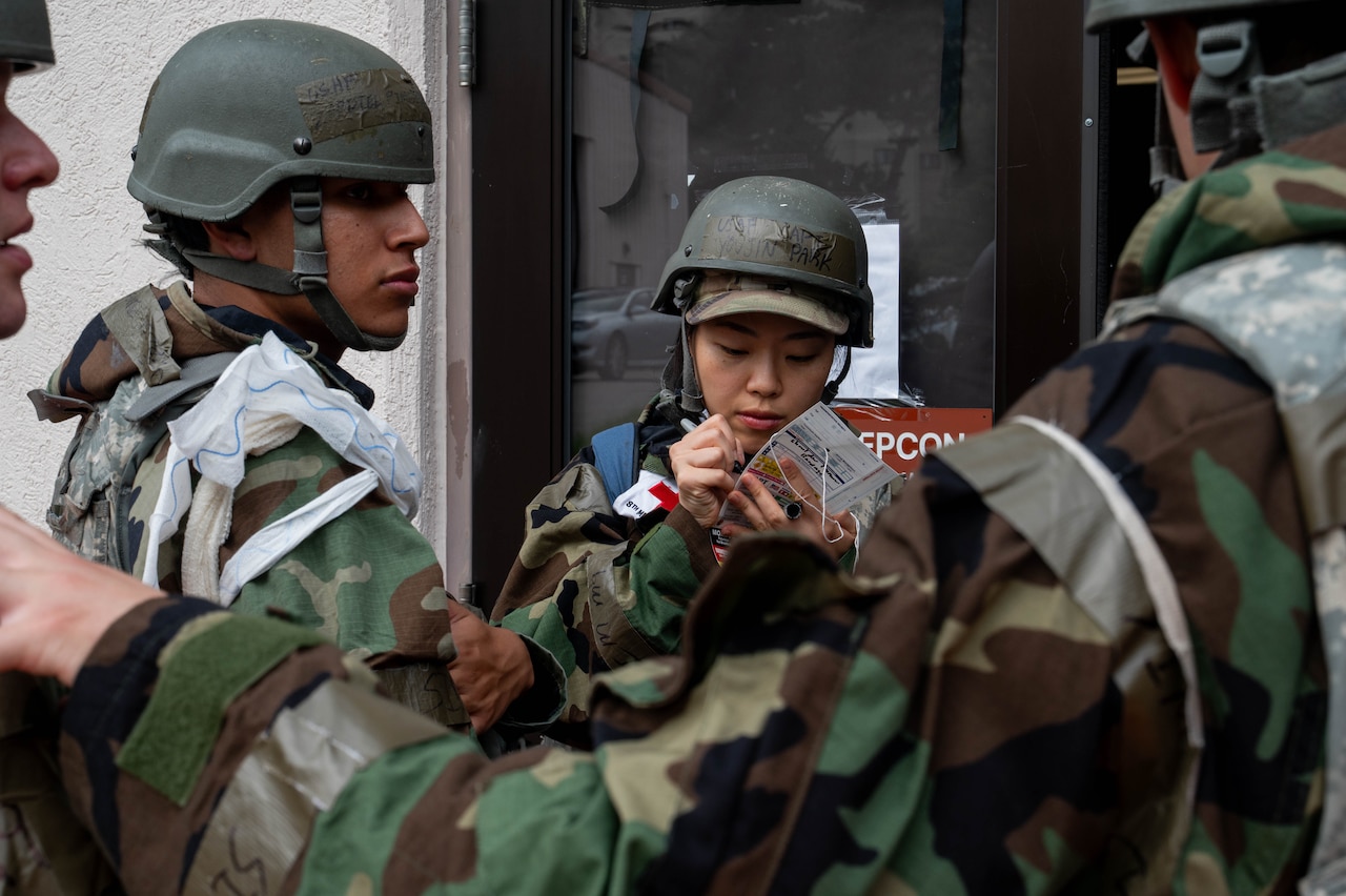 A woman in a camouflage military uniform and helmet writes on a piece of paper while standing outside a building; there are three people in similar attire standing in front of her.