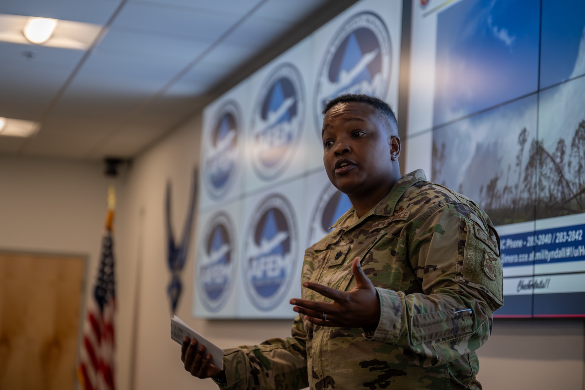 A close-up of a female Airman in a camouflage uniform speaking and gesturing during a presentation, with a video wall and an American flag in the background.