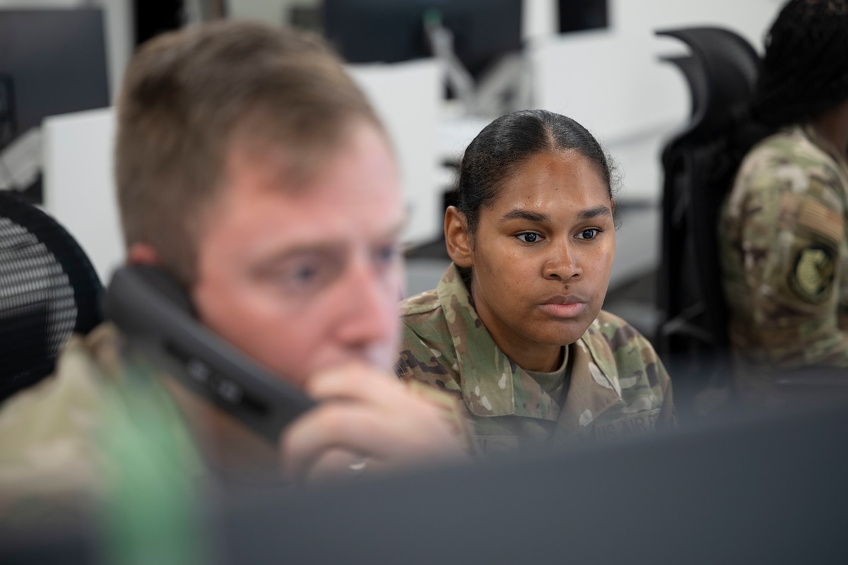A uniformed Airman with her hair in a bun looks intently at her computer screen in an office, while a man in the foreground is on the phone.